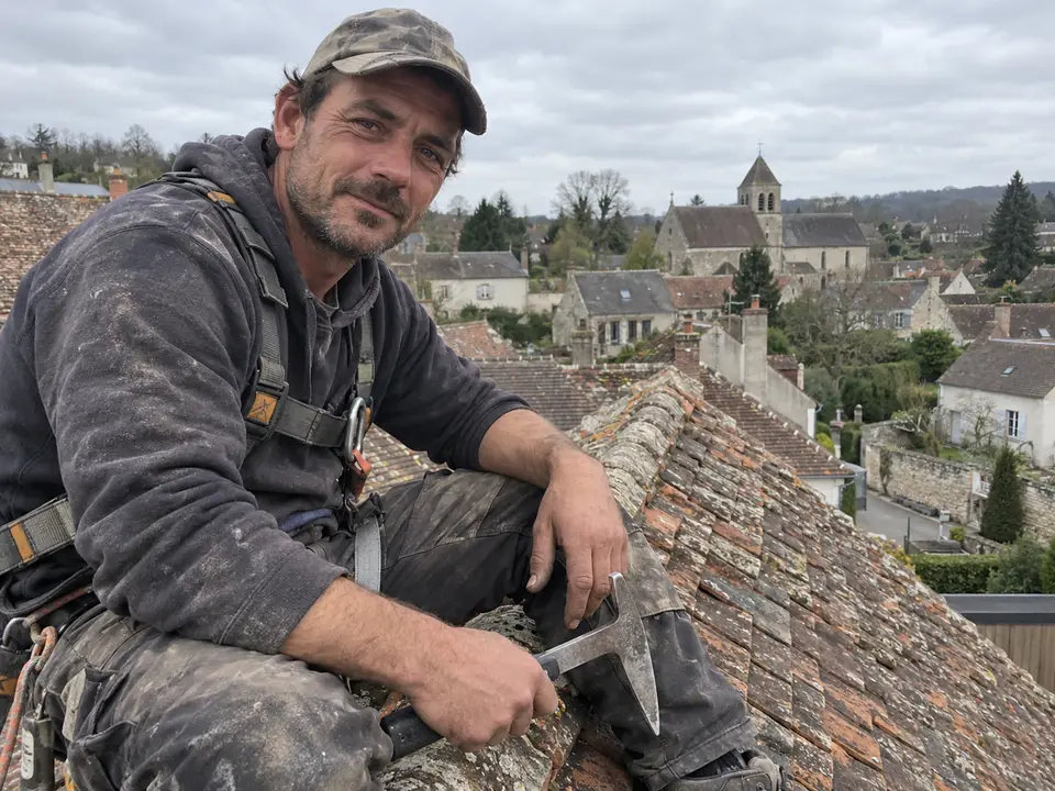 Julien Bastide, couvreur, assis sur le faîtage d'un toit en tuiles plates, marteau à la main